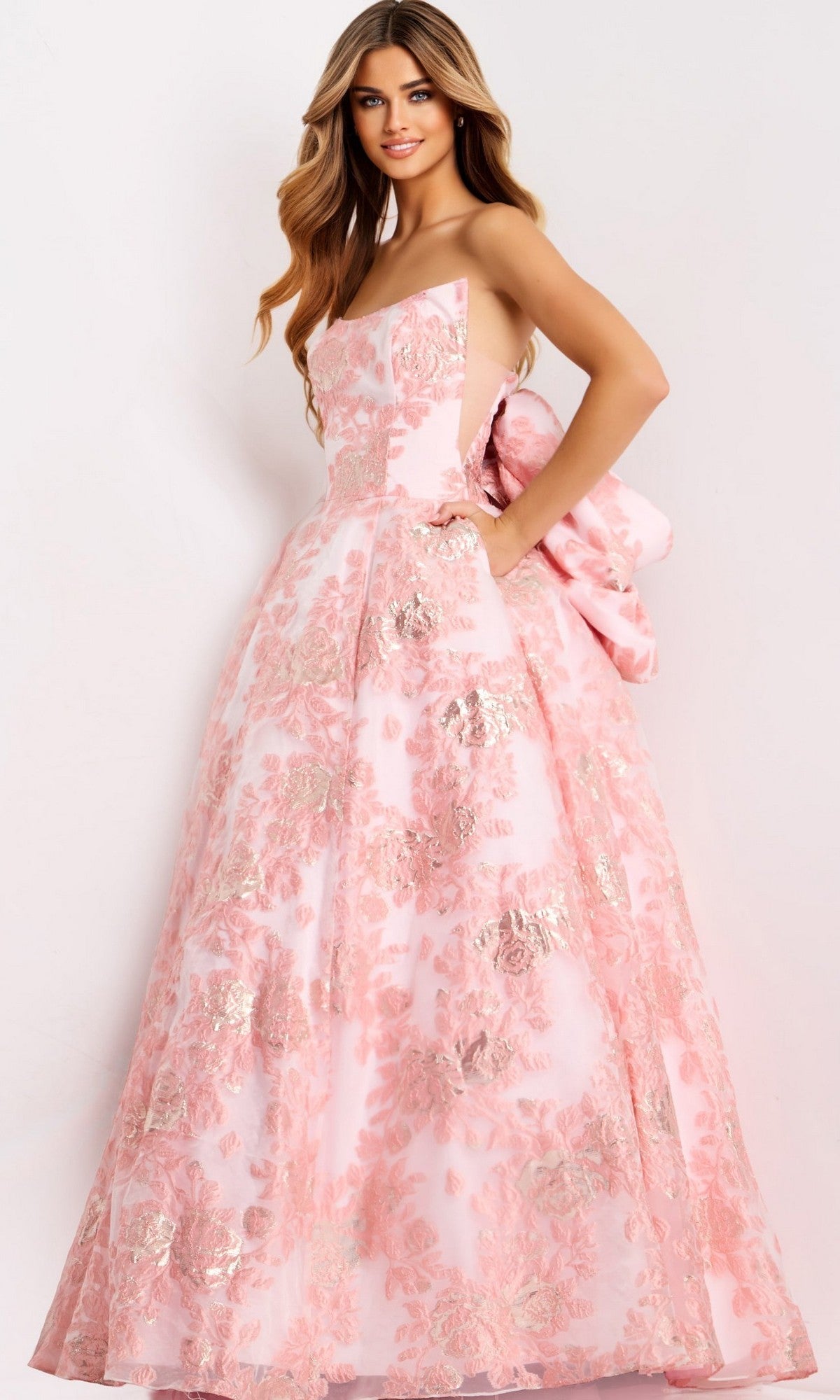 A woman models the Jovani 47702 long prom dress—a strapless jacquard ball gown with metallic floral patterns and a large back bow—posing against a plain light background.